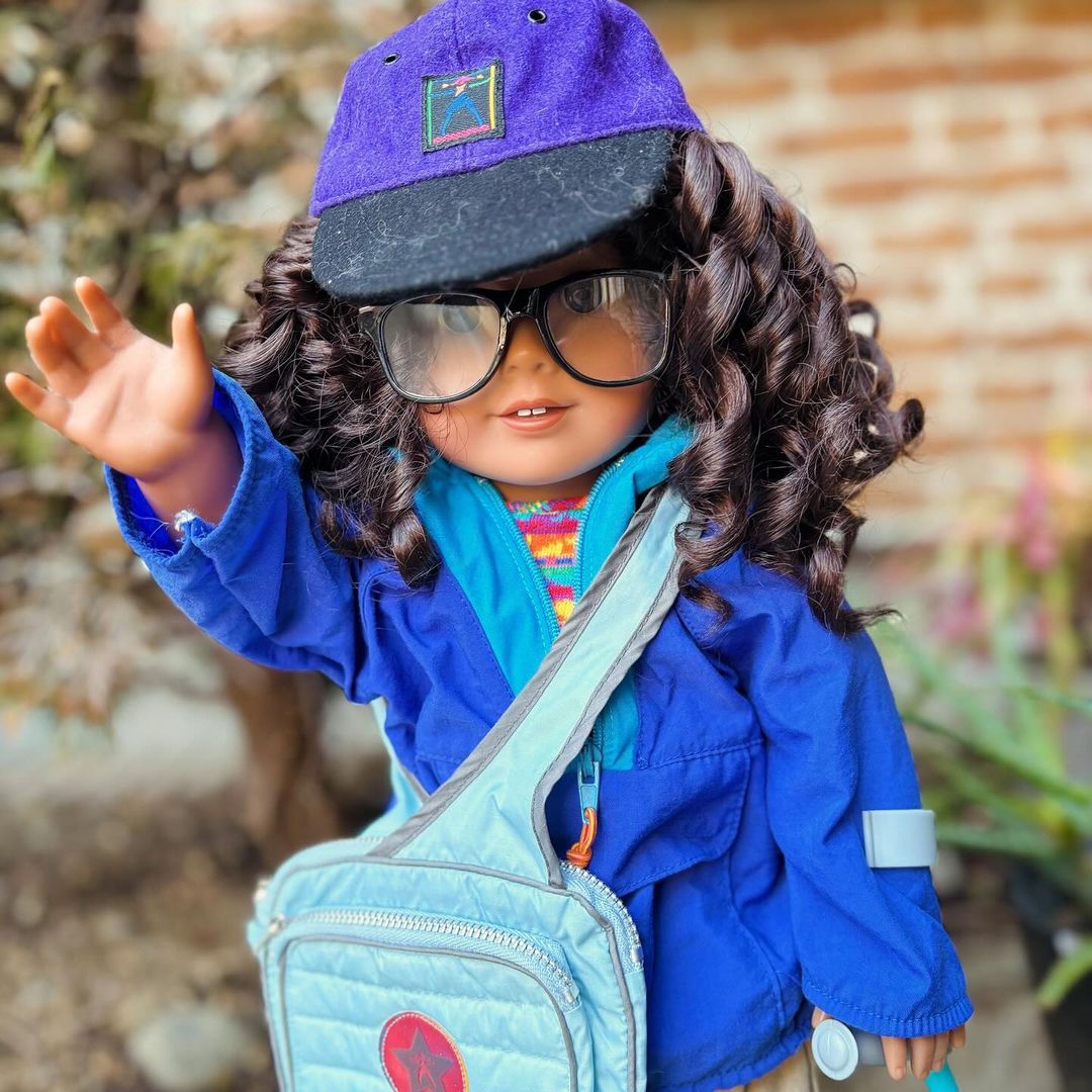 tan American Girl with curly dark brown hair and gray eyes wearing a blue windbreaker; she has glasses and a cane